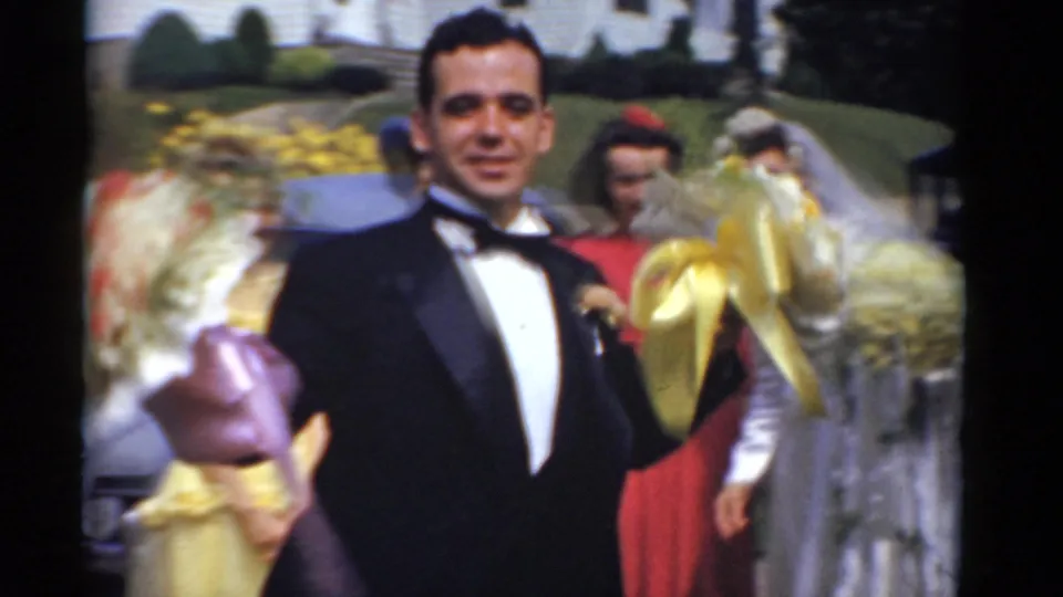 The groom in a tuxedo faces the camera as bridesmaids with bouquets stand close around him in the outdoor wedding group.