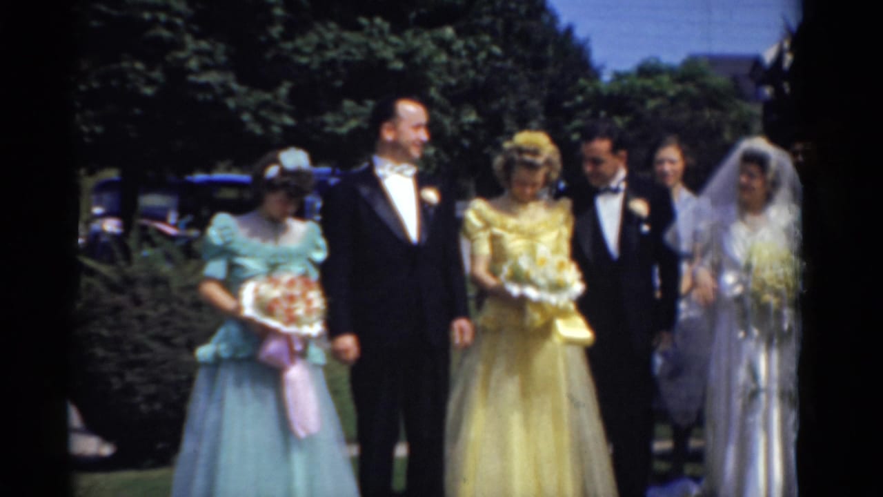 Wedding party outdoors with bride, groom, bridesmaids, tuxedoed men, bouquets, trees, and a car behind them.