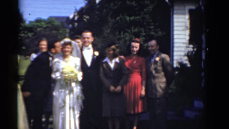 Outdoor 1975 Bridgeport wedding scene with the bride, groom, and guests grouped near greenery and a residential-looking structure.
