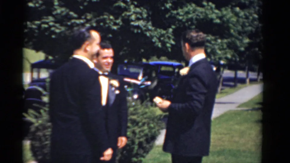Bridgeport, Connecticut 1975 groomsmen in dark tuxedos laugh and talk near a classic car in a sunny yard.