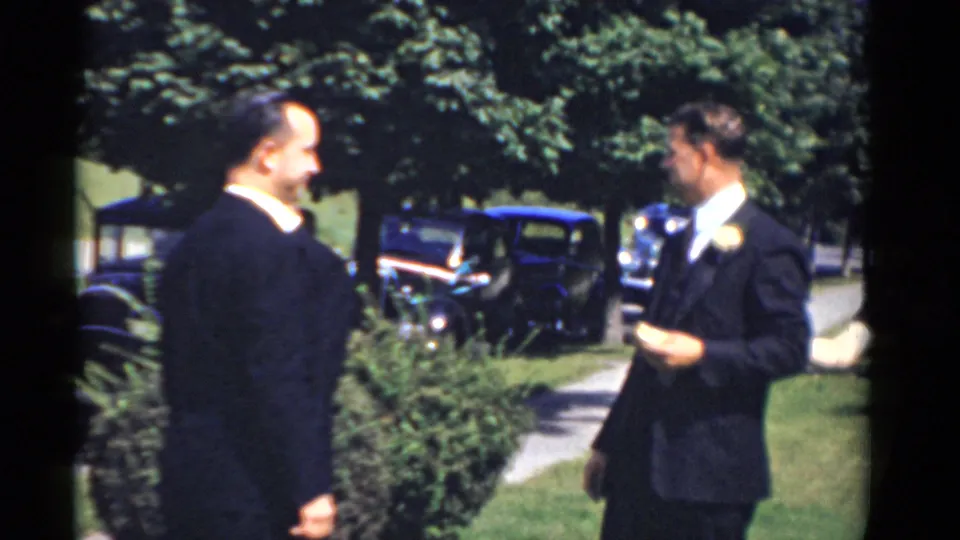 Two men in dark formalwear converse outdoors in Bridgeport, Connecticut, with lawn, trees, sidewalk, and 1970s-era cars behind them.