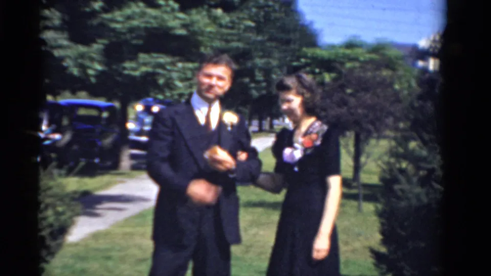 A man in a dark suit with a boutonniere stands with a woman in a dark dress during a 1975 Bridgeport outdoor celebration scene.