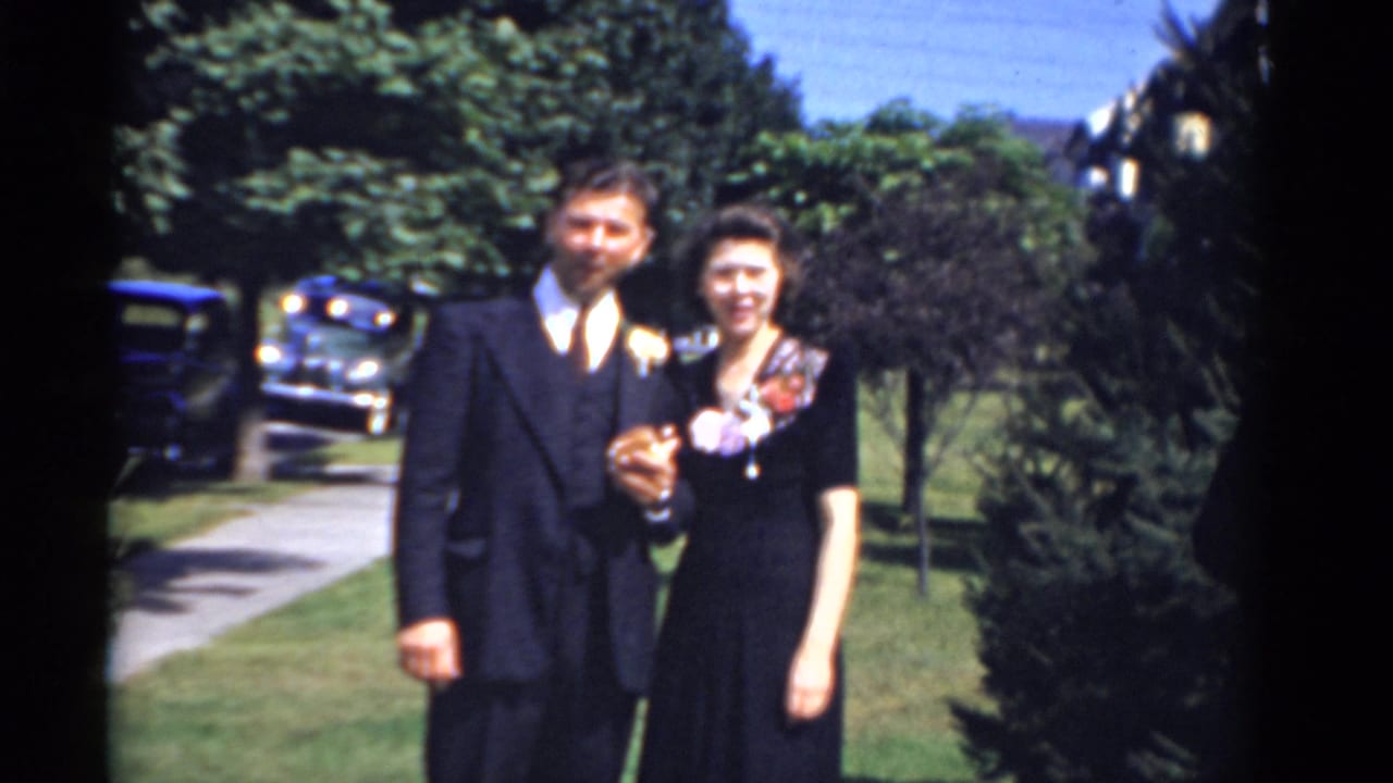 Man in a dark suit and woman in a black dress pose together on a sunny lawn.