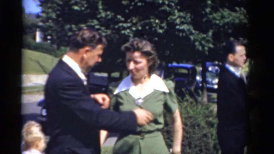 The 1975 home-movie frame shows a suited man and a woman in a green dress interacting on a lawn, with another guest partly visible nearby.