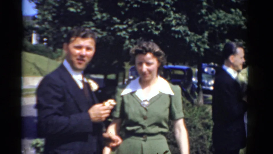 1975 Bridgeport wedding footage shows the groom and a woman in green standing near a street with older cars and trees in the background.
