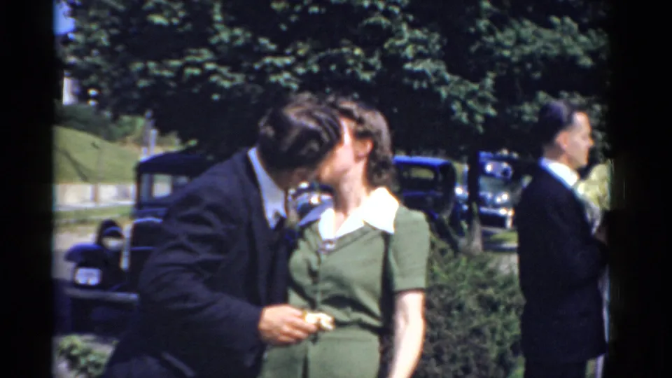 Outdoor 1975 Bridgeport wedding frame with a groom leaning toward a woman in green for a kiss near parked cars and trees.