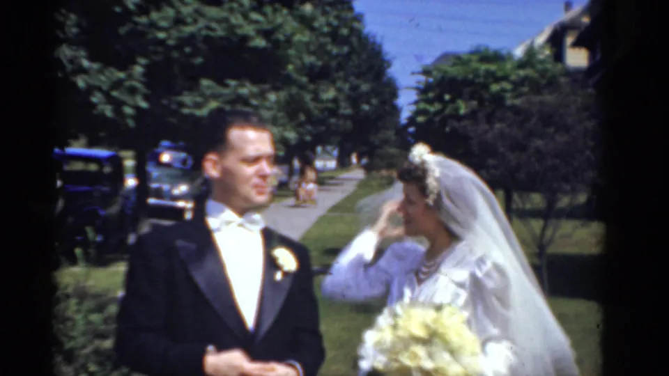 1975 Bridgeport wedding frame showing bride in white veil and bouquet beside groom in dark formalwear with trees and cars behind them.