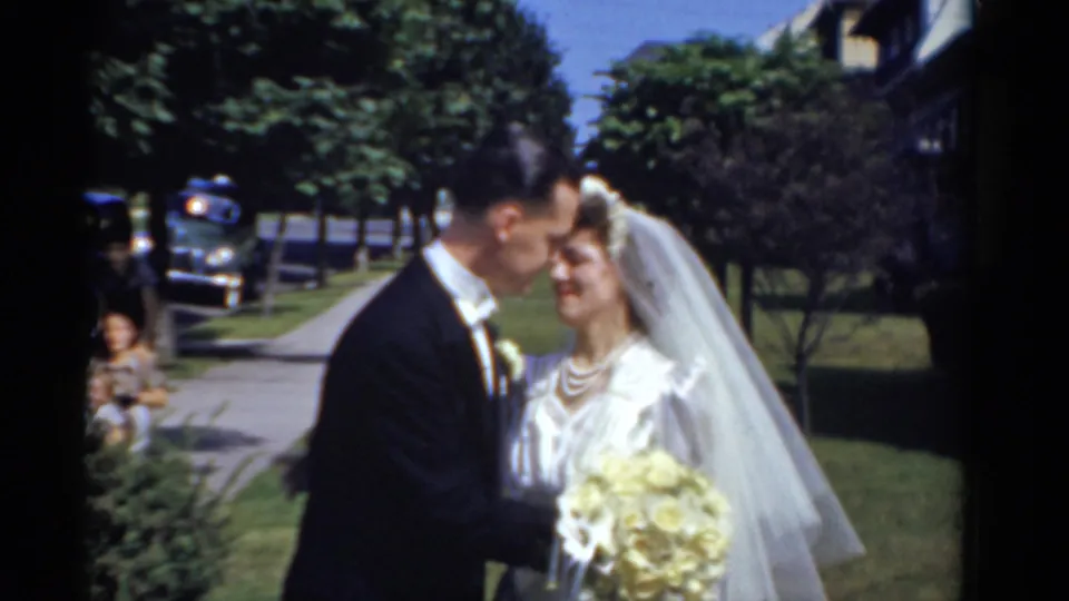 In 1975 Bridgeport, Connecticut, a bride in veil and groom in black tuxedo pose closely on a sunny residential street.