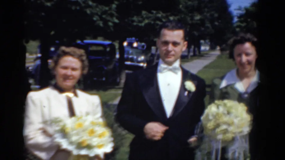 A groom poses with two women outdoors in Bridgeport wedding footage, with older cars visible along the residential street.