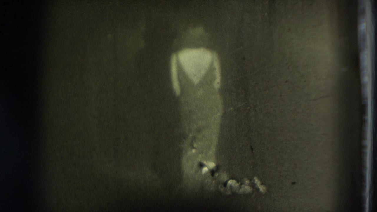 Faint figure in light clothing appears in a dim 1975 San Francisco 8mm home-movie frame with heavy grain and soft focus.