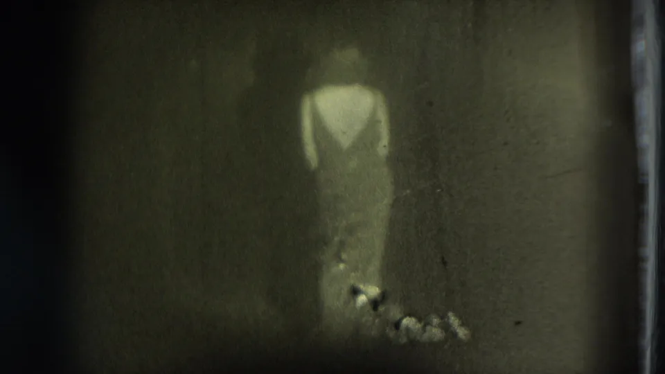 Faint figure in light clothing appears in a dim 1975 San Francisco 8mm home-movie frame with heavy grain and soft focus.