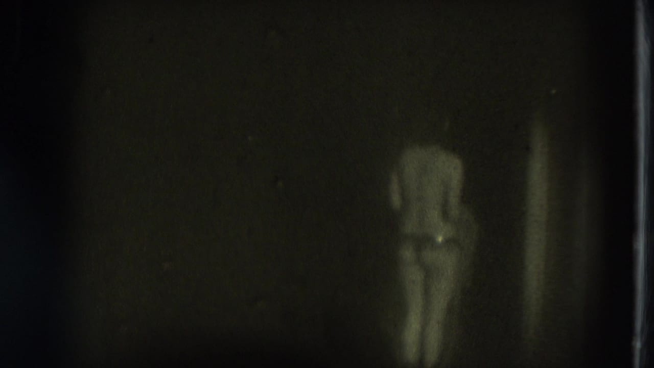 Adult woman in minimal clothing stands in a dark, grainy room.
