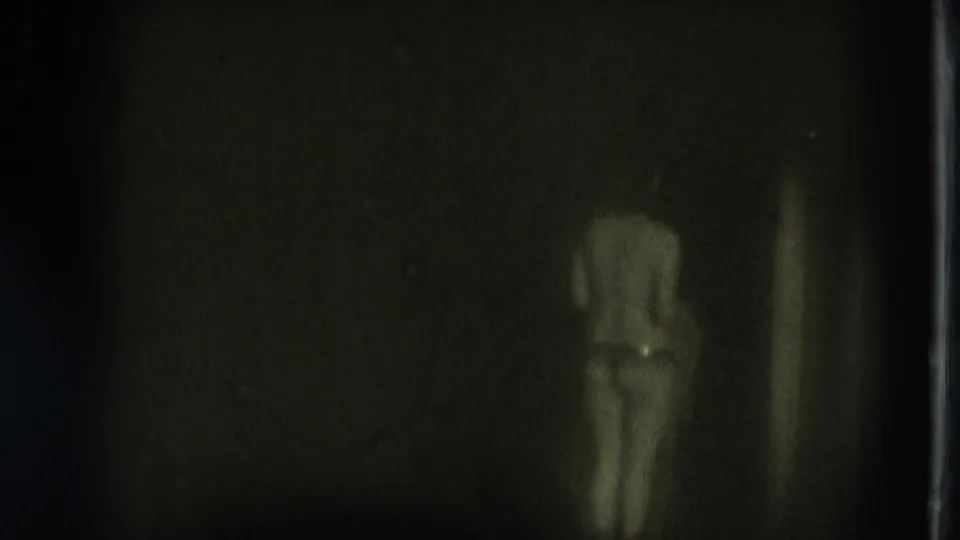 1975 San Francisco 8mm frame showing an adult woman in minimal clothing seen from behind in a dim, green-tinted room.
