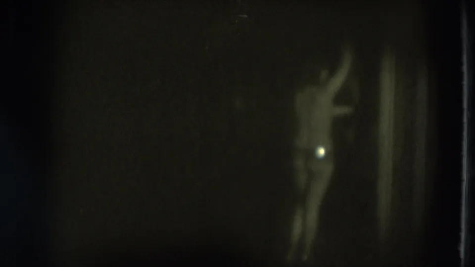 1975 San Francisco 8mm frame of an adult woman dancing in a dim room, with an arm raised and a small light flare nearby.