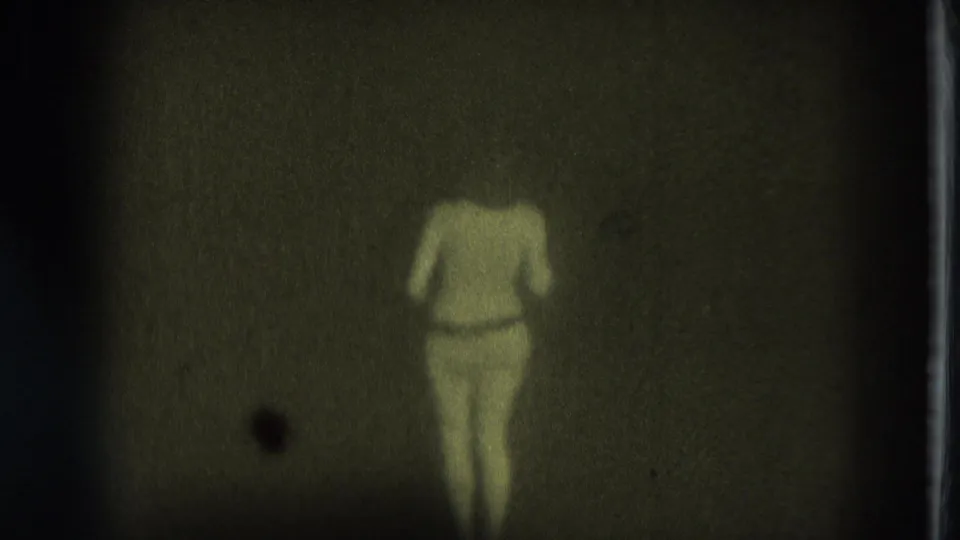 1975 San Francisco 8mm frame of an adult woman in minimal clothing dancing within a brighter patch of an otherwise dark room.
