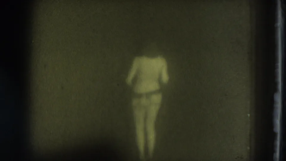 1975 San Francisco home-movie still of an adult dancer in soft focus, turned away in a dark indoor space.