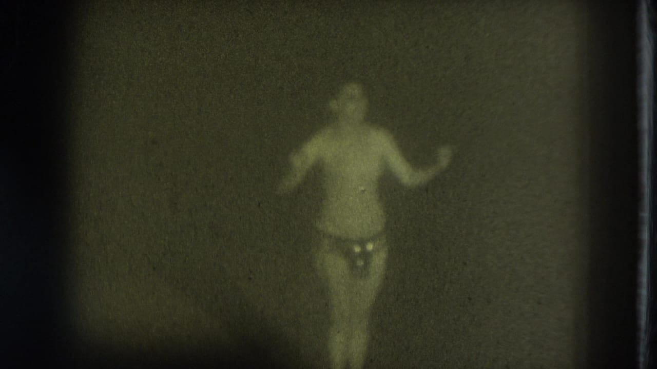Soft-focused performer in minimal stage attire standing in a dark space with one arm raised.
