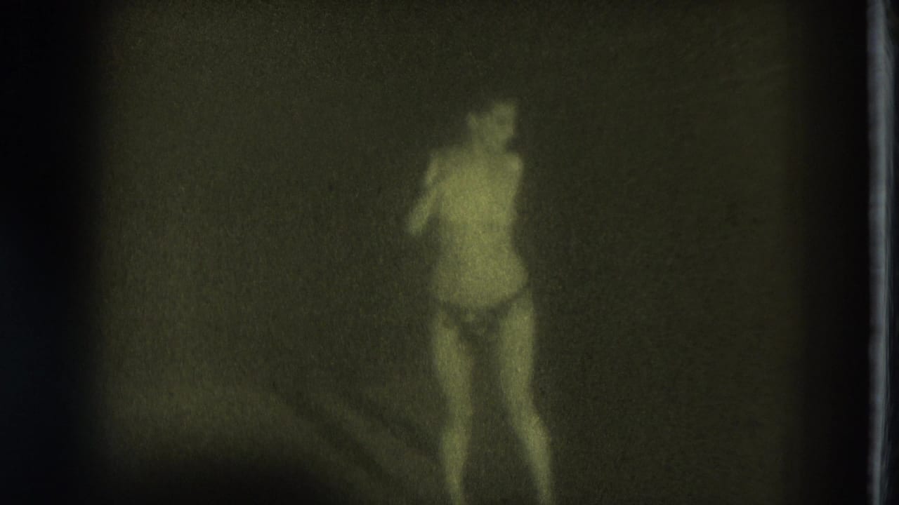Minimally clothed woman standing on a dark stage in soft green-tinted 8mm footage.
