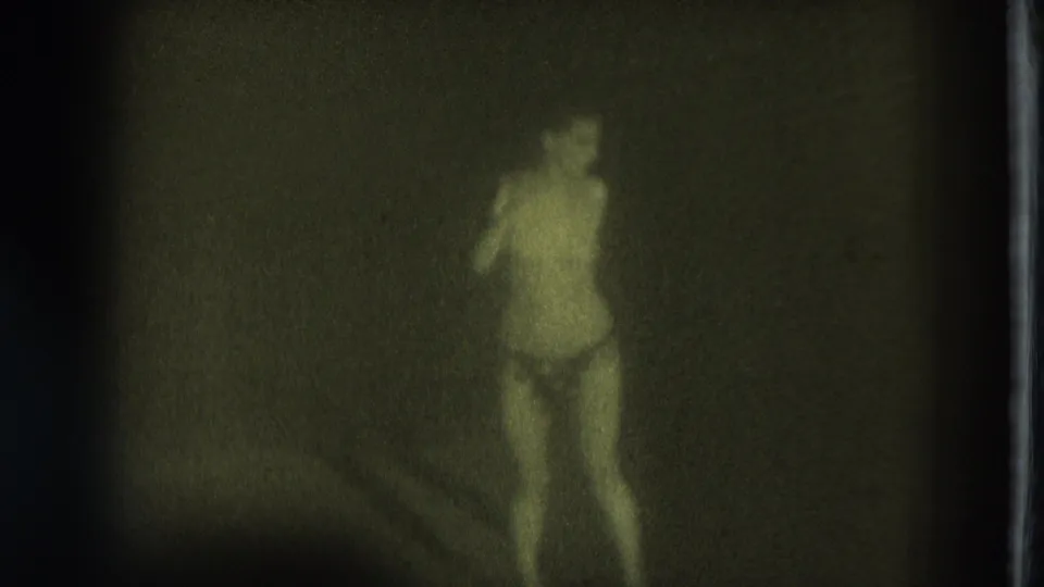 1975 San Francisco 8mm frame showing a minimally clothed female performer standing in dim stage light with heavy grain.