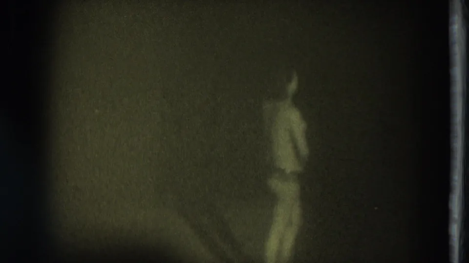 1975 San Francisco 8mm frame with a minimally clothed performer in profile against a dark, grainy stage background.