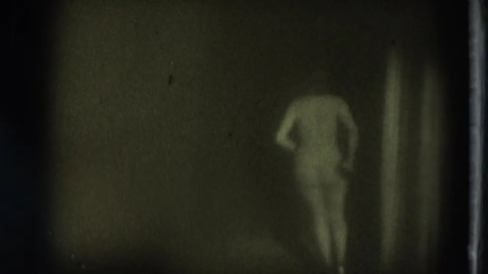 1975 San Francisco 8mm frame showing the performer from behind near a vertical edge in a dim, green-tinted stage setting.