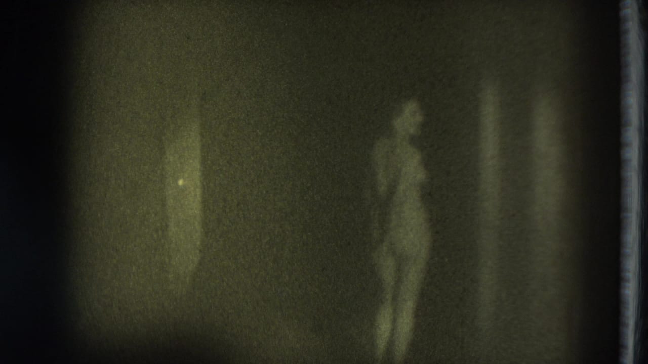 Soft, dark frame showing a nude adult woman standing or beginning to move in a dim interior.