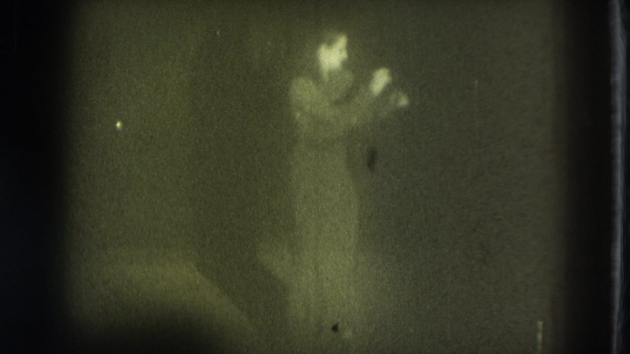 1975 San Francisco 8mm frame showing a distant, blurred stage performer under dim light with strong grain and green-yellow cast.