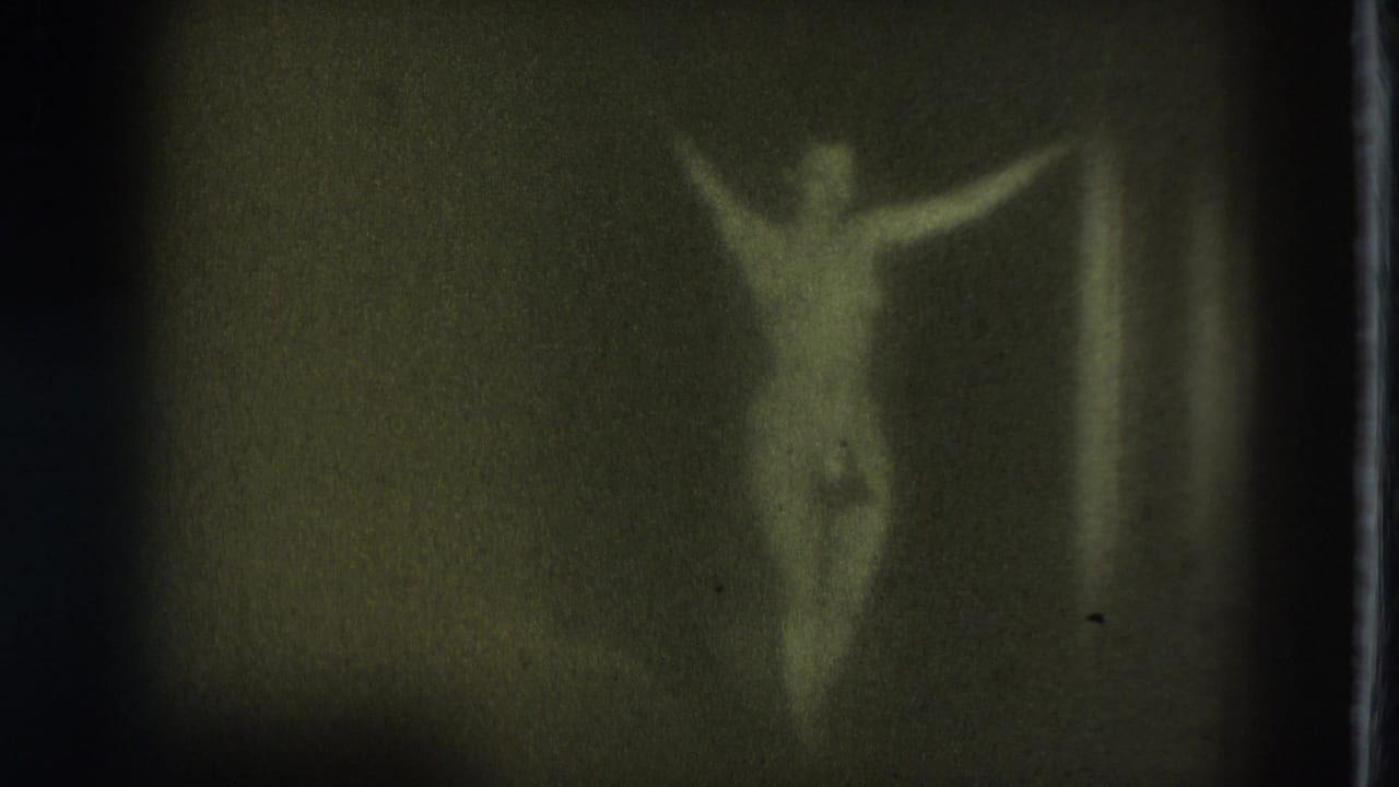 Topless adult performer in a g-string stands on a dim stage with arms raised.