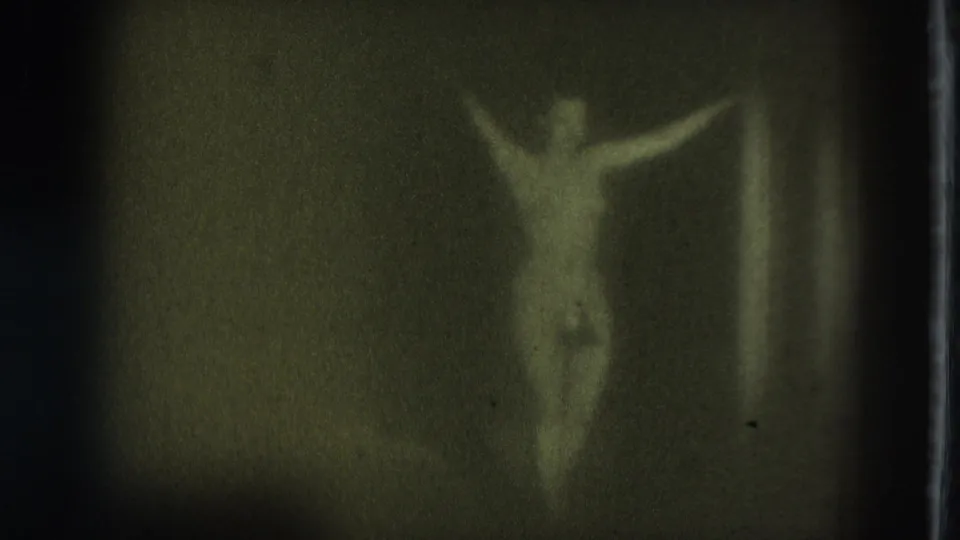 In metadata-supported 1975 San Francisco footage, a topless adult performer appears faintly lit on a dark stage with arms extended.