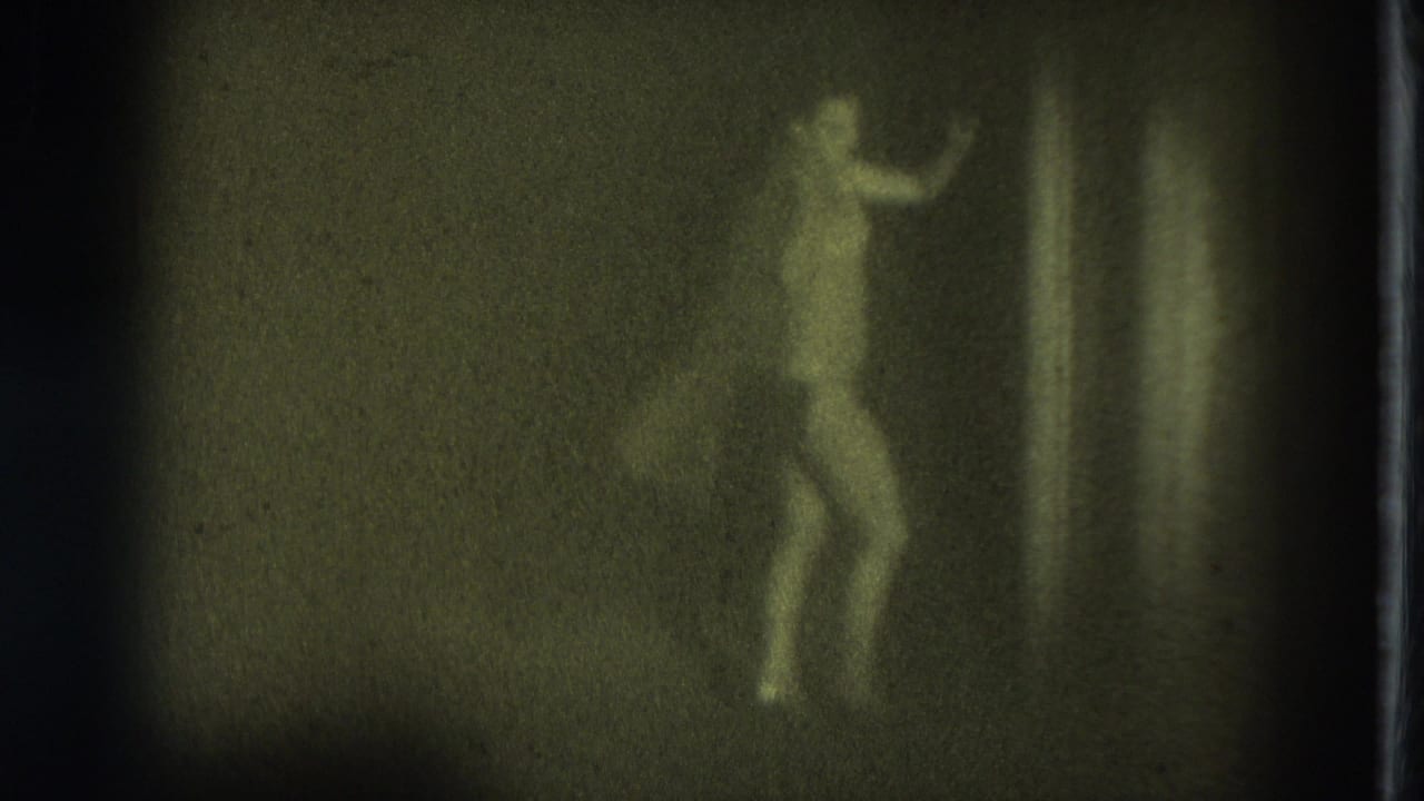 Topless adult performer moves sideways near the right side of a dark stage.