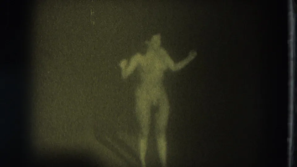A blurred adult performer in a g-string appears partly turned on the dark stage, with one arm extended in the 1975 San Francisco footage.