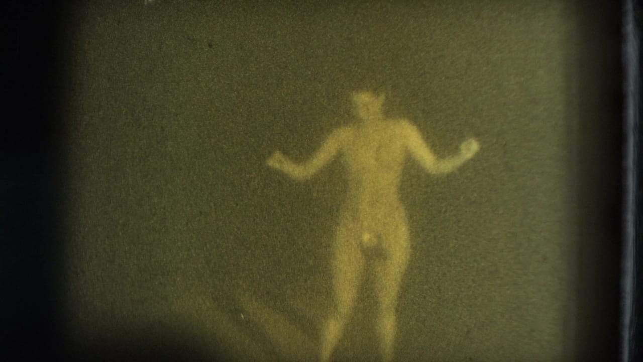In supplied 1975 San Francisco 8mm footage, a nude adult woman appears through soft grain with arms lifted while dancing or posing.