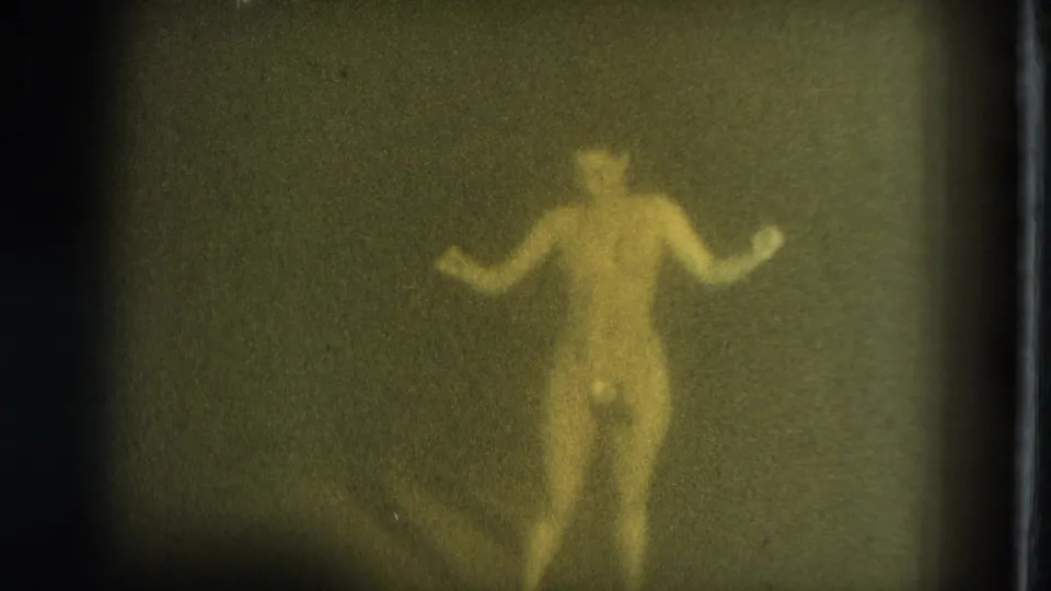 In supplied 1975 San Francisco 8mm footage, a nude adult woman appears through soft grain with arms lifted while dancing or posing.