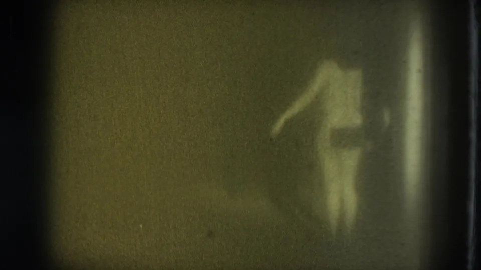 A dim 8mm frame shows a nude adult woman partially obscured by shadow or translucent material, with one arm extended.