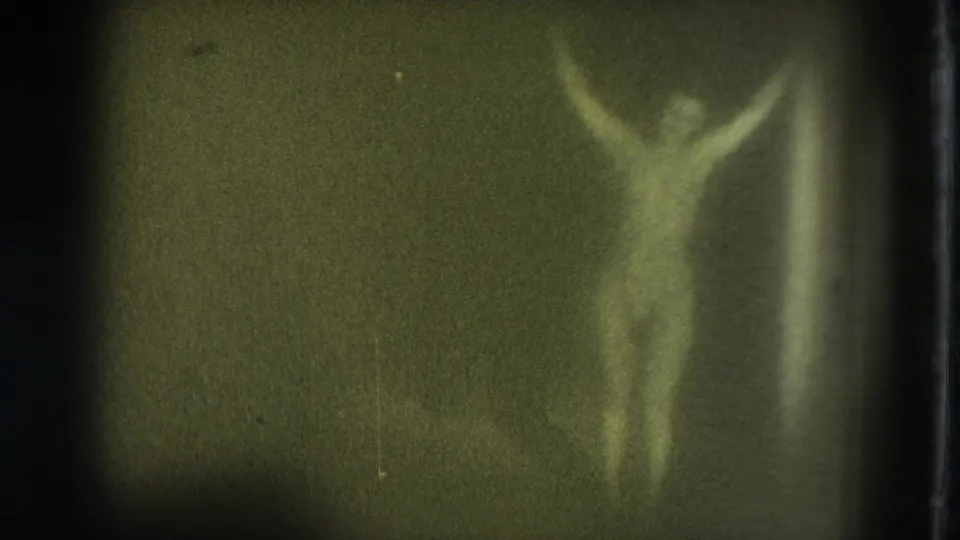 The 1975 8mm frame shows a nude adult woman in soft focus, arms lifted overhead against a dark, yellow-green background.