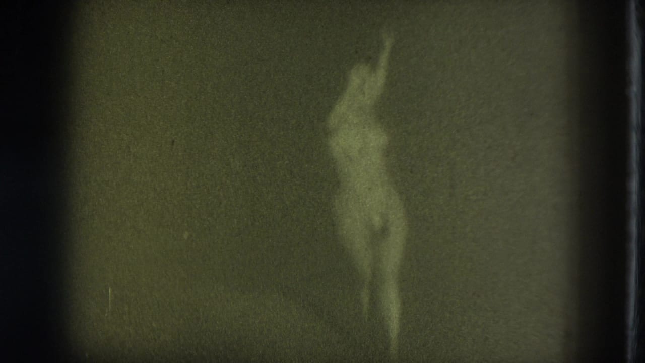 Soft, grainy frame of an adult woman wearing pasties with one arm raised against a dark backdrop.