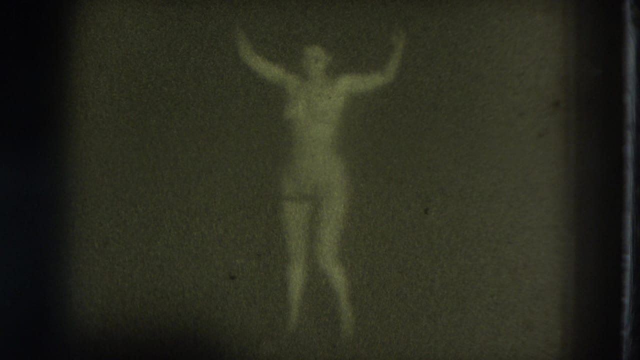 1975 San Francisco 8mm frame of an adult woman wearing pasties with both arms raised while dancing in low light.