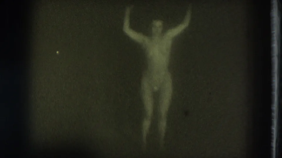 1975 San Francisco 8mm frame of an adult woman wearing pasties, arms bent upward as she dances in dim light.