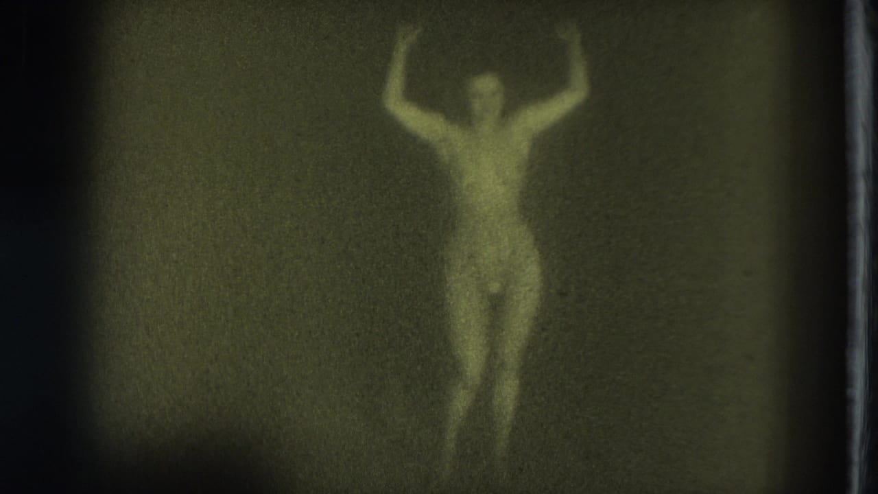 Green-tinted 1975 San Francisco 8mm frame of a nude adult woman standing and dancing with arms raised in a dim interior.