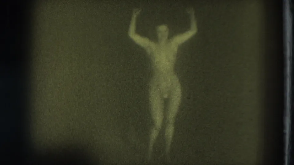 Green-tinted 1975 San Francisco 8mm frame of a nude adult woman standing and dancing with arms raised in a dim interior.