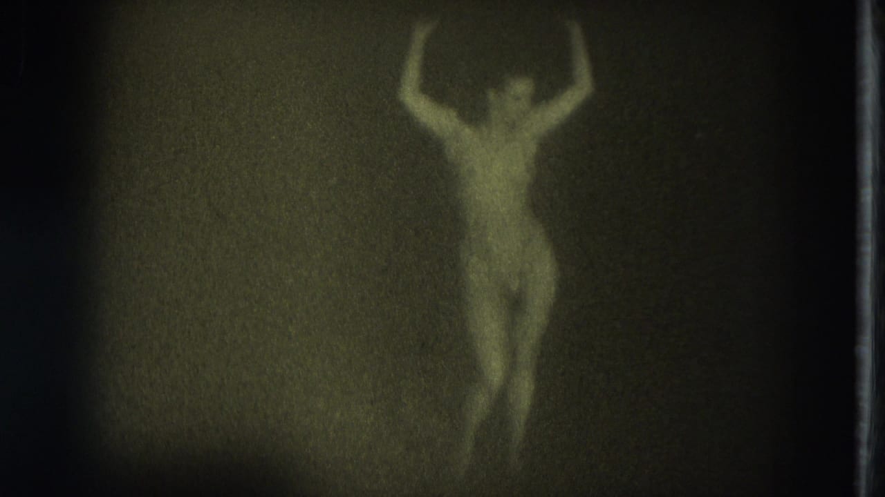 Green-yellow 8mm frame from the supplied 1975 San Francisco clip showing a nude adult woman posed with arms raised in low light.