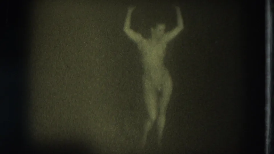 Green-yellow 8mm frame from the supplied 1975 San Francisco clip showing a nude adult woman posed with arms raised in low light.