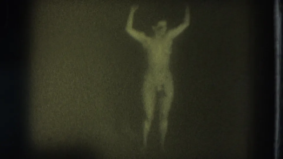 Soft, grainy 8mm frame of the adult dancer with arms raised and body turned slightly in a dim green-tinted interior.