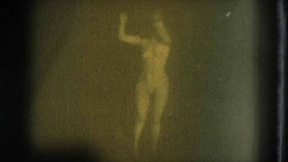 1975 8mm frame of a nude adult woman in soft focus, standing front-facing in dim light against a dark backdrop.