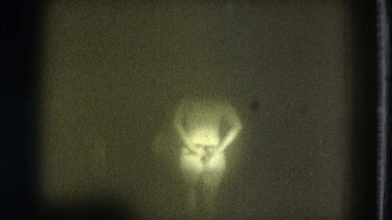 Soft, yellow-tinted 8mm frame showing a nude adult figure bent forward against a dark, indistinct background.