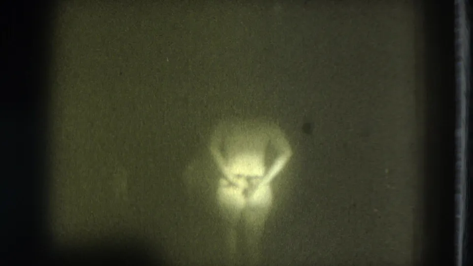 Soft, yellow-tinted 8mm frame showing a nude adult figure bent forward against a dark, indistinct background.