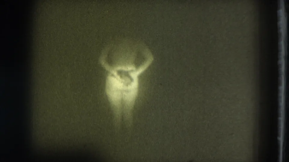 1975 8mm frame of a nude adult figure in dim light, body softly outlined with arms raised or bent near the shoulders.
