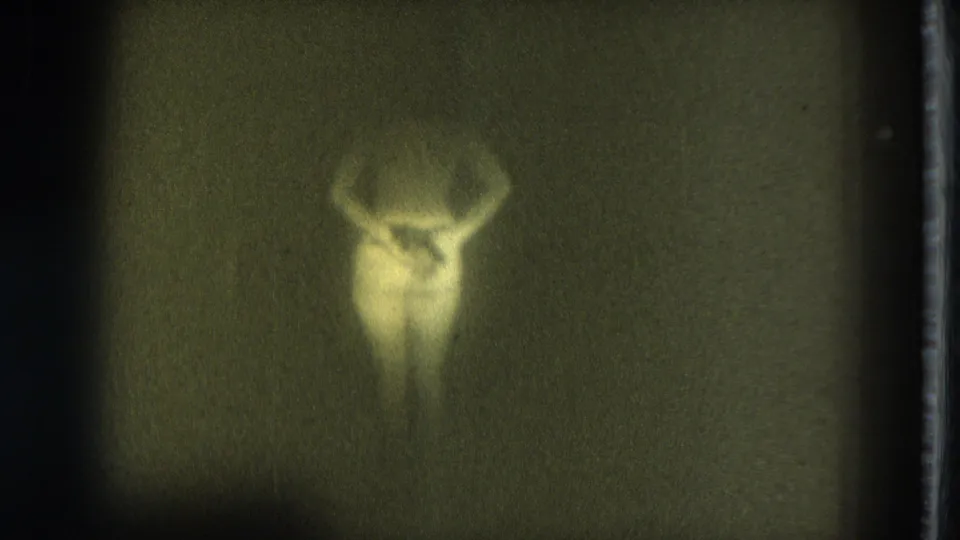Soft 8mm frame from 1975 showing a nude adult figure turned away or partly side-on, lit faintly against a dark background.