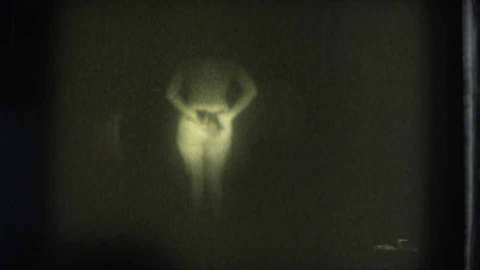 Low-light 1975 8mm frame from San Francisco showing a blurred woman lowering her arms against a dark background.
