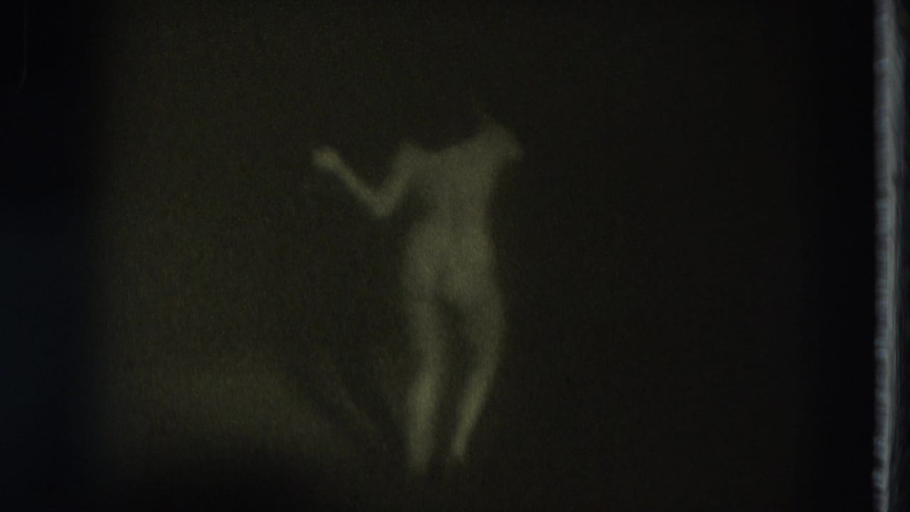 Green-tinted 1975 8mm frame from San Francisco showing a nude adult woman in soft focus with one arm raised against a dark interior wall.